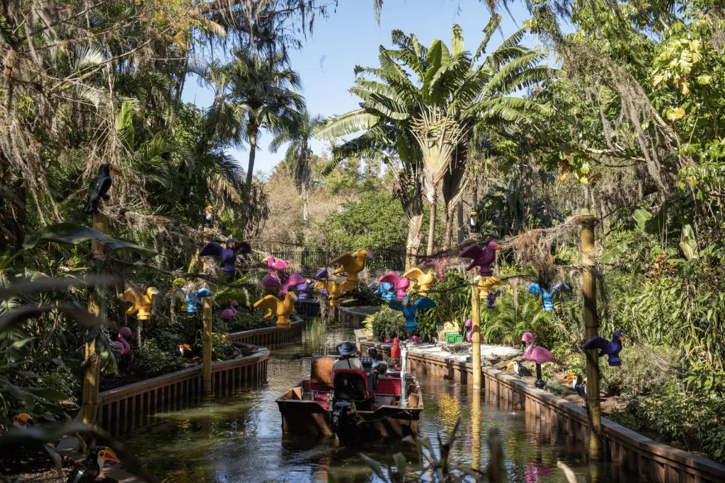 A boat sailing through a waterway lined by tall trees and greenery, with LEGO birds overhead