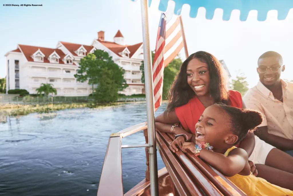 A mother, father and young girl looking out at the water on a boat. A white, Victorian-style building is in the background. 
