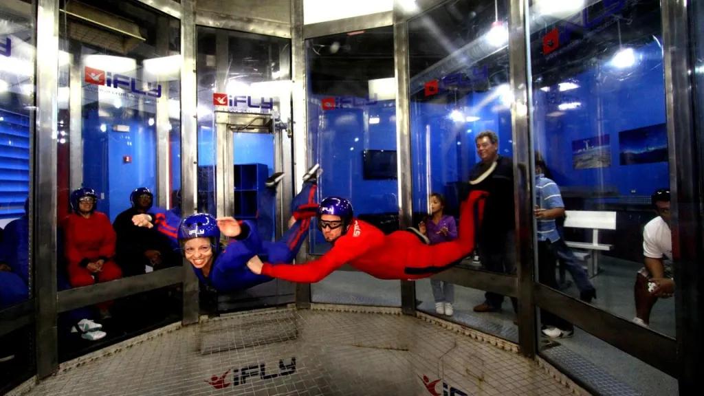 A large fan with windows all around inside a blue building and people peering in watching as a man in a full red jumpsuit, a helmet and goggles holds onto a girl wearing a blue jumpsuit, a helmet and goggles as the both float in starfish positions.