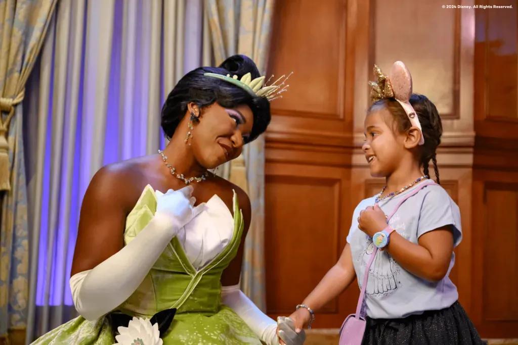 Ein junges Mädchen mit rosa Minnie Mouse Ohren hält Händchen mit Prinzessin Tiana 