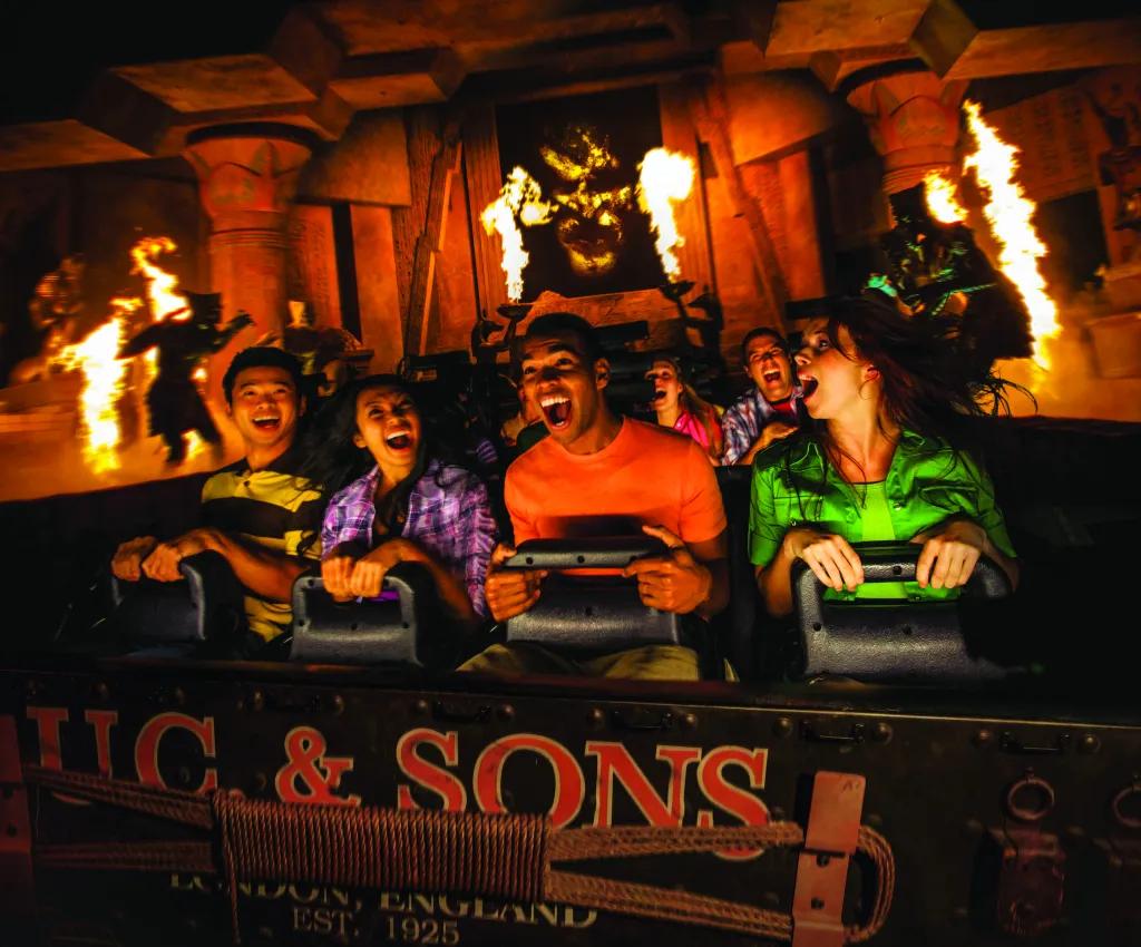 Four guests smiling and gasping as they clutch to their lap bars on a indoor rollercoaster. There is fire pyrotechnics surrounding them and silhouettes of mummy's jumping out on them.