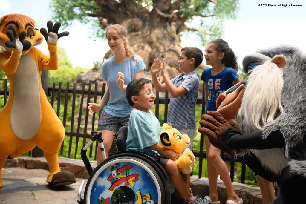 A young boy in a wheelchair smiling at Rafiki, a cartoon monkey from the Lion King movie. There are three young kids behind them smiling looking at Timone, a cartoon meerkat from the Lion King. Behind there is a large tree with carvings of different animals on the large stump.