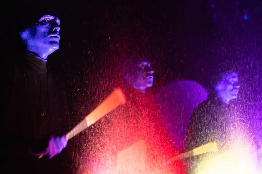 Three men painted completely blue wearing black outfits are holding glowing sticks and smacking a drum which has loads of neon paint of different colours.