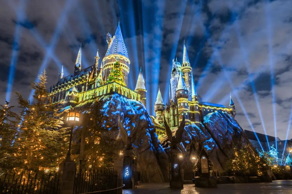 Hogwarts castle lit up in projections of Christmas decorations and Christmas trees. There are also spotlights projected in the night sky.