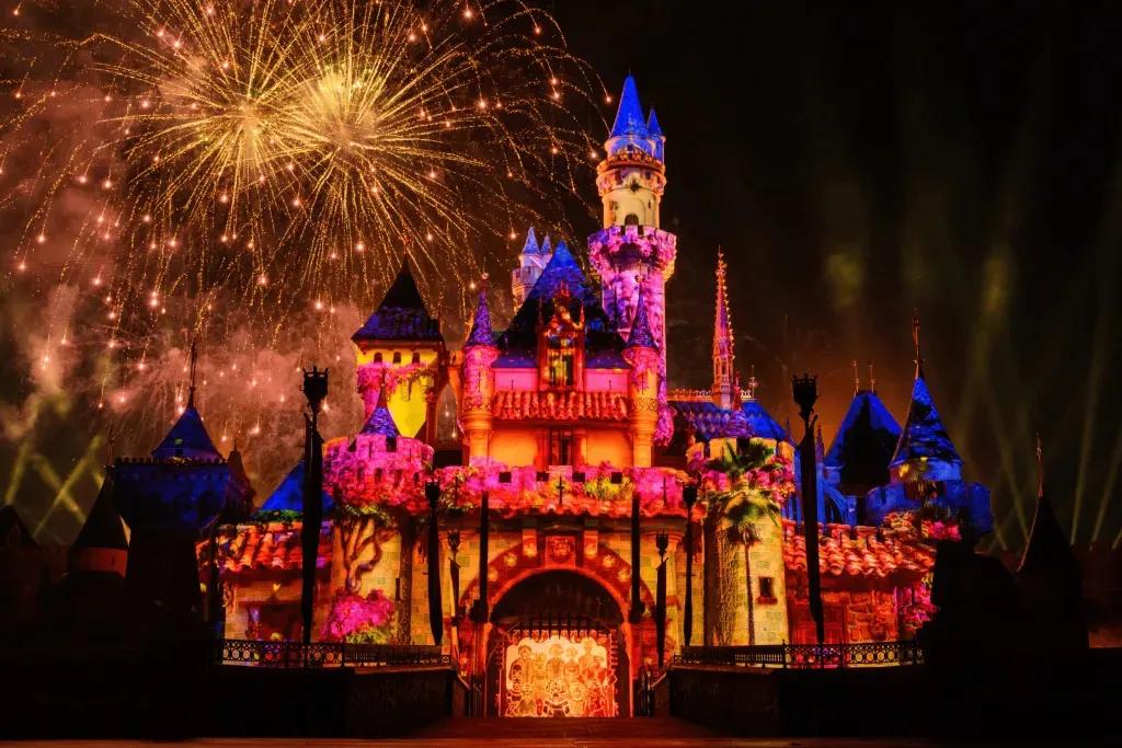Das kleine rosafarbene Schloss mit den blauen Dächern wird mit einer Projektion von Encanto beleuchtet und in der Mitte steht die Familie Madrigal in einem Familienporträt zusammen. Dahinter füllt ein gelbes Feuerwerk den Himmel.