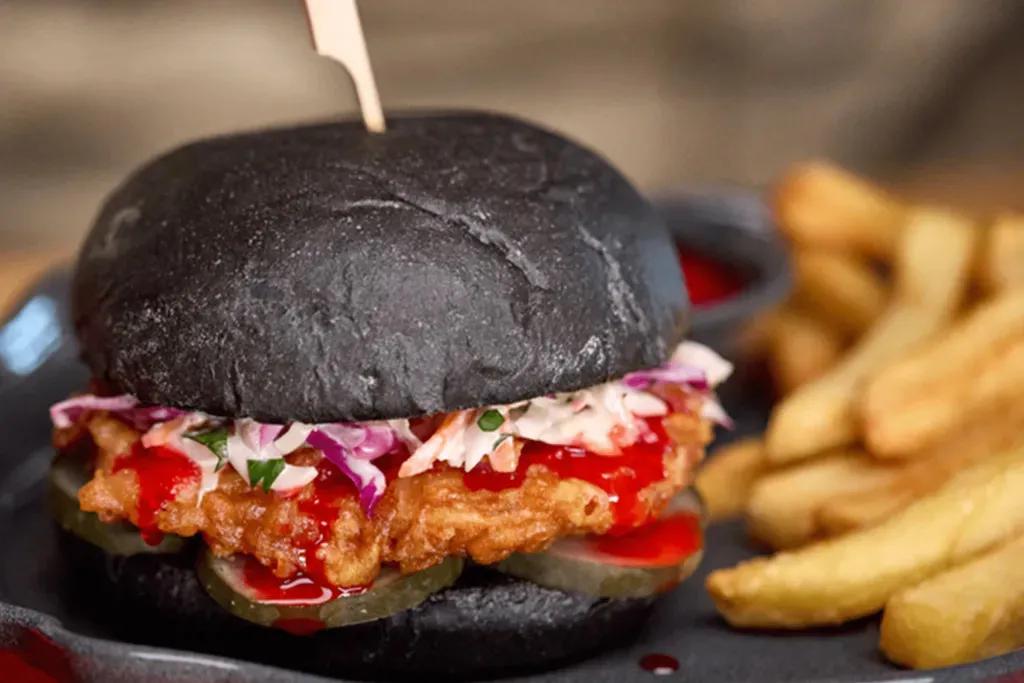 Schwarzes Burgerbrötchen mit gebratenem Hähnchen, Krautsalat, Zwiebeln, Essiggurken und einer leuchtend roten Soße. Dazu gibt es Pommes und einen roten Dip.
