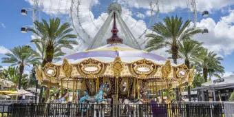 A gold and white carousel with different pastel coloured horses and gold bars, in front of 6 palm trees diagonally lined behind and a huge ferris wheel which is white.