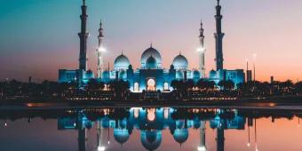 A beautiful religious temple in Abu Dhabi located overlooking a lake. The sun is setting with a pink and blue hue in the sky and the temple is perfectly reflected in the lake. It is a white building with three white domes and four big pillars sticking out.