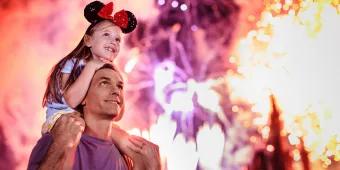 Young girl sat on her dads shoulders as the both look into the sky watching the magnificent fireworks above Cinderella Castle. There are many different colours and lights.