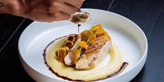 Chicken or fish dish on top of smooth mash potato with green plant on top and crackers. There is a mans hand pouting dark sauce around the dish on the clean white bowl.