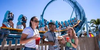 Three people stood in front of Pipeline, a standing up rollercoaster, holding small plates and cups full of red cocktails.