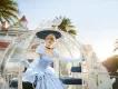 Cinderella, a blonde princess in a blue ball gown is stepping out of a glass carriage in front of a clean large white hotel.