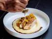 Chicken or fish dish on top of smooth mash potato with green plant on top and crackers. There is a mans hand pouting dark sauce around the dish on the clean white bowl.