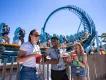 Three people stood in front of Pipeline, a standing up rollercoaster, holding small plates and cups full of red cocktails.