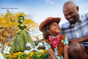 A young boy dressed as a cowboy smiling up at his dad. They are both sat in front of a topiary shaped like Bo Peep from Toy Story