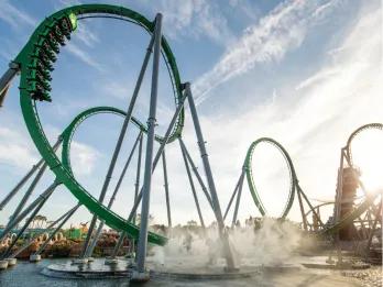 Giant green rollercoaster with two vertical loops and a car in motion on track diving towards lake surrounded by mist