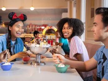 Three adults laughing while sat at a table eating a large ice cream sundae