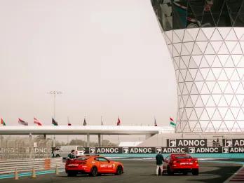 Two red cars on race track with two men stood next to either one. There is a white structure in the background with black windows and then flags lining the track.