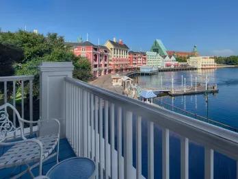 There is a balcony with a white chair and a small table overlooking a lake. Alongside the building there are different coloured buildings, red, yellow and green.