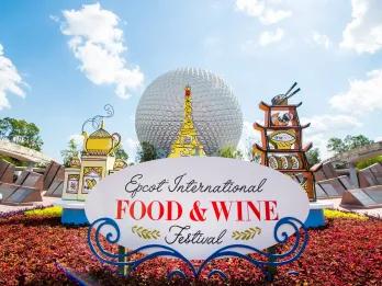 White circular building shaped like a golf ball with a plant pot in front with a sign saying "Epcot International Food and Wine Festival" with cardboard cutouts of international recognisable structures such as the Eiffel Tower, built out of food.