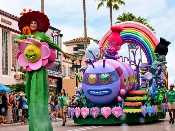 Woman on stilts dressed like a flower with a face on it with a huge hat on. Behind her pink troll called Poppy riding a caterpillar creature on a float is waving at crowds.