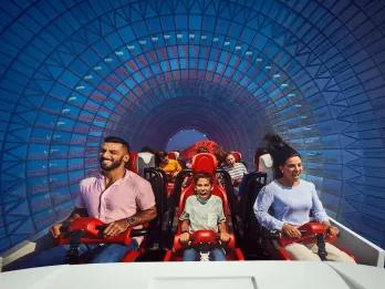 A row of three guests sat on a white roller coaster car with red lap bars holding them in. There is a tunnel surrounding coloured blue and they are all screaming with excitement.