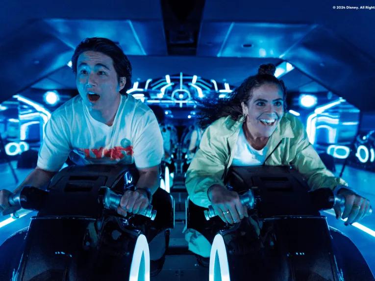 Two young adults sat on motorcycles shaped vehicles TRON Lightcycle. They are leaning forward gasping with shock. 